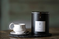 White ceramic cup and saucer with a black cylindrical container on a wooden surface.