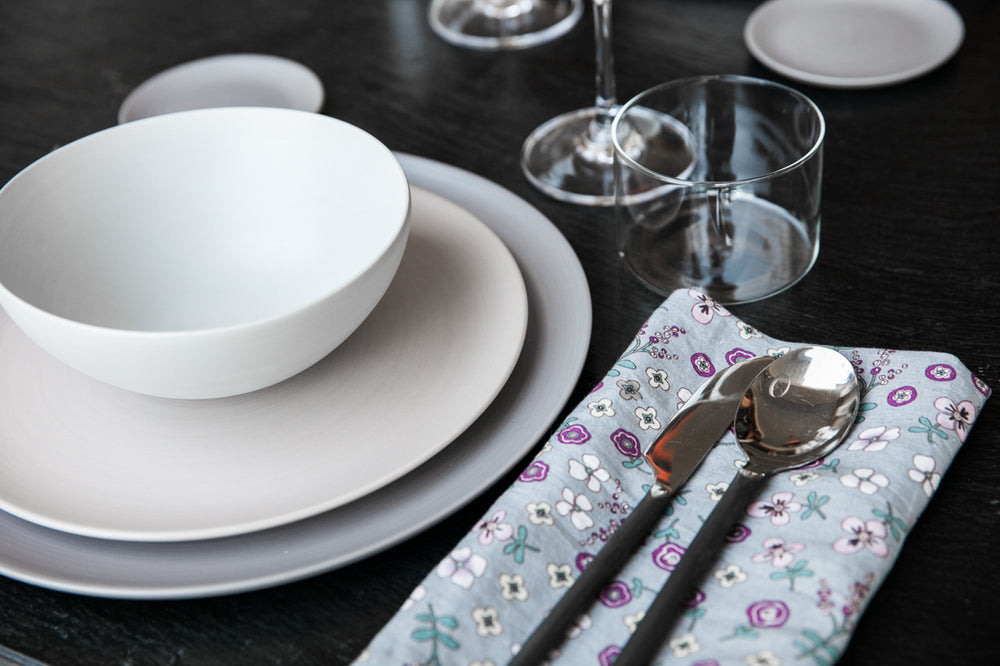 A set of handmade ceramic dishes including a dinner plate, side plate, coaster, side bowls, and a serving bowl, displayed on a table with cutlery and glasses.